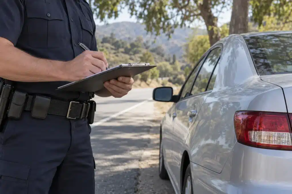An officer writing on a clipboard beside a parked silver sedan